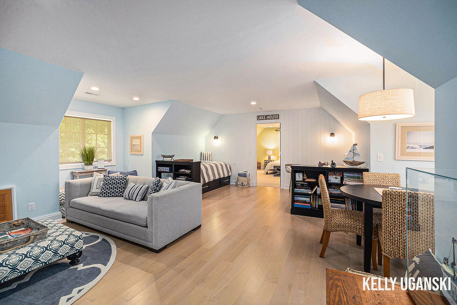 This is a spacious guest bedroom featuring a seating area with a gray sofa and patterned ottoman, a sleeping area with a twin bed, and a dining area with a round table and wicker chairs. The room is painted in a light blue hue, complemented by light wood flooring and natural light from a window. The overall impression is a cozy and versatile space.