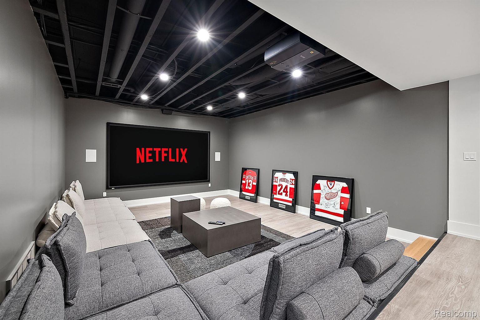 This is a modern living room featuring a large sectional sofa in gray and beige tones, a dark gray coffee table, and a large screen displaying the Netflix logo. The room has a dark ceiling with recessed lighting and gray walls, complemented by framed sports jerseys on the wall. The overall impression is a comfortable and stylish entertainment space.