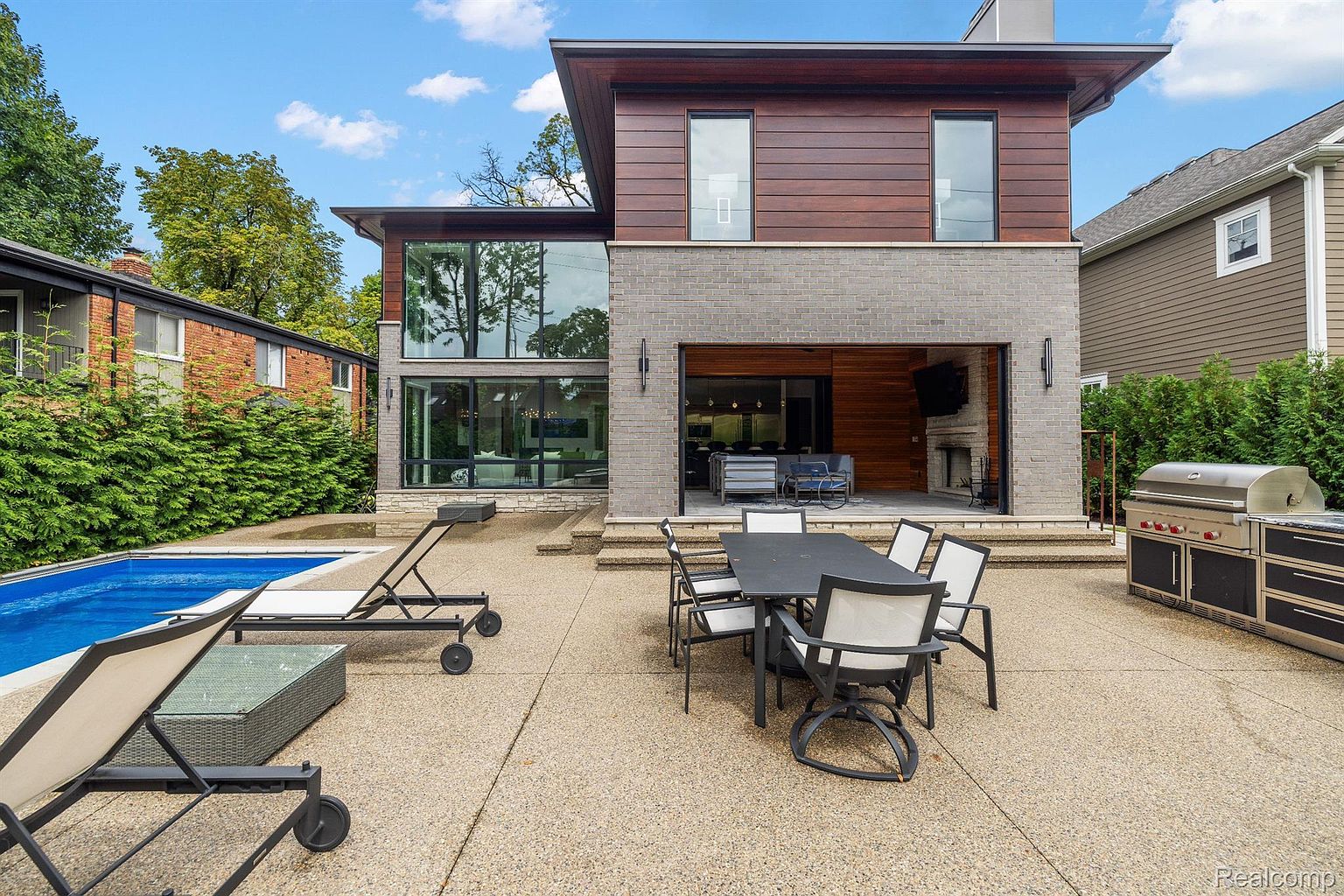 This image showcases the rear exterior of a modern home, featuring a blend of wood and brick facade. A spacious patio includes outdoor dining furniture and a grill, extending from an open indoor/outdoor living space. A swimming pool is visible to the side, complemented by lounge chairs, creating an inviting backyard oasis.