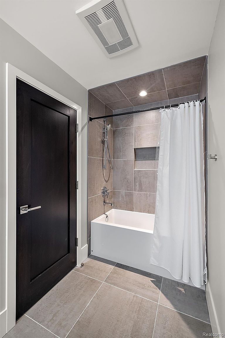 This is a well-lit bathroom featuring a modern design. The bathroom includes a bathtub with a shower, enclosed by a shower curtain and tiled walls. A dark-colored door with a silver handle is visible on the left, and the floor is covered with large, gray tiles.