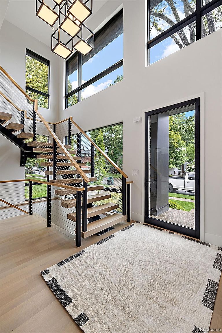 This interior shot showcases a modern entryway with a striking staircase featuring floating wooden steps and sleek metal railings. Natural light floods the space through large windows, highlighting the light wood flooring and a contemporary area rug. A black-framed glass door provides a view to the exterior, enhancing the open and airy feel of the home.