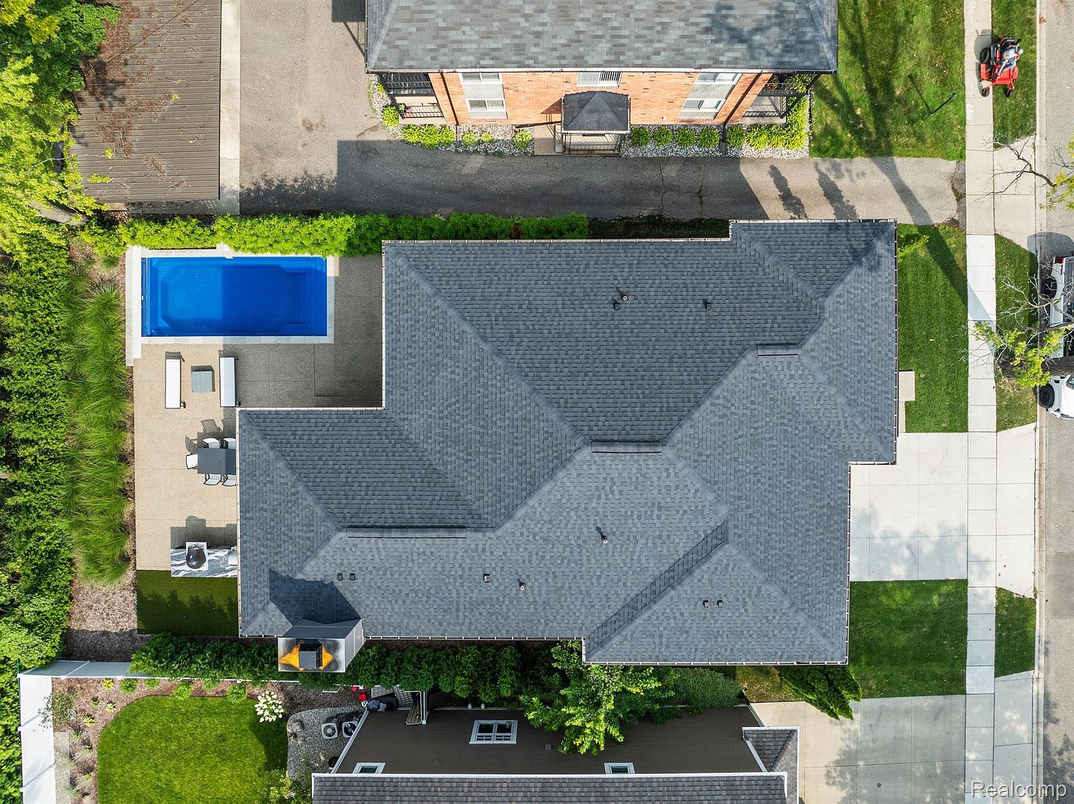 This aerial shot showcases a well-maintained property featuring a large house with a dark gray roof, a rectangular swimming pool, and a patio area with outdoor furniture. The surrounding landscape includes lush greenery, manicured lawns, and neighboring buildings, creating a serene and private setting. The perspective offers a comprehensive view of the property's layout and its integration with the surrounding environment.