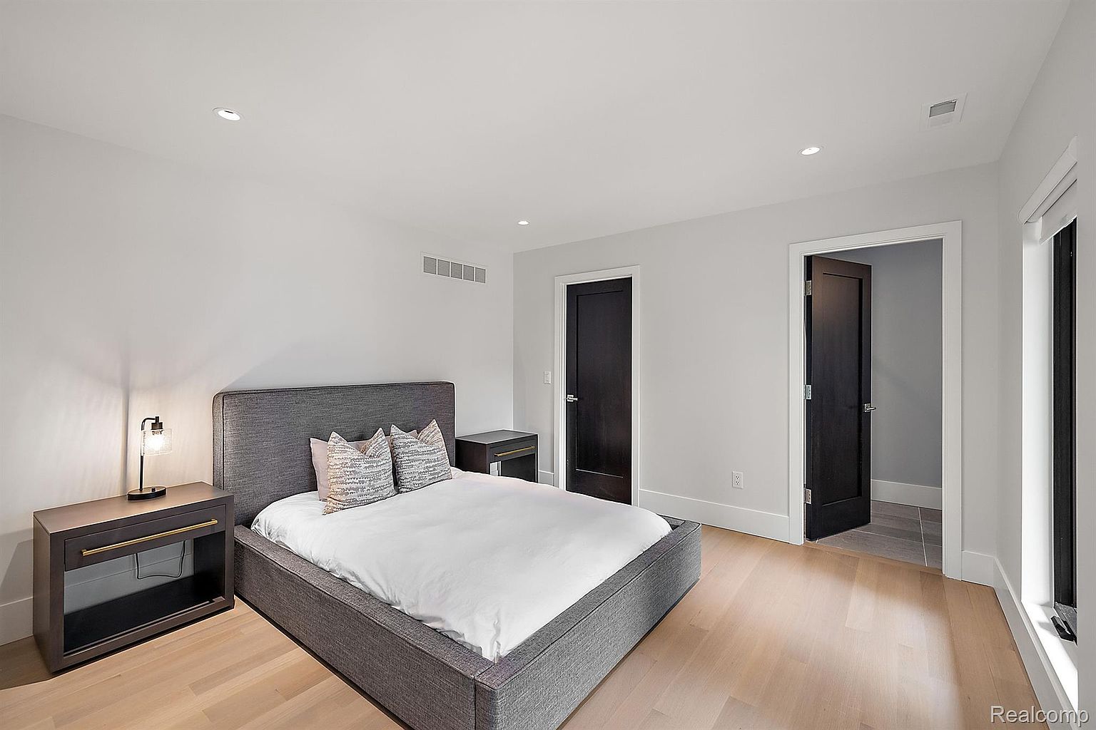 This is a well-lit primary bedroom featuring a gray upholstered bed with white linens and decorative pillows. The room has light hardwood floors, white walls, and dark-colored doors, creating a modern and minimalist aesthetic. Two dark wood nightstands flank the bed, and recessed lighting provides ample illumination.