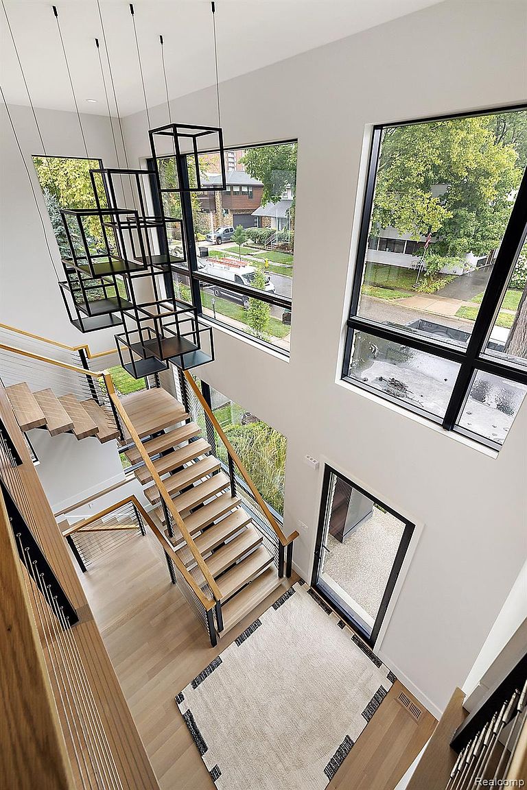 This is an interior shot of a modern home's hallway and staircase, viewed from an elevated angle. The staircase features light wood steps with metal and wood railings, complemented by a unique, geometric chandelier. Large windows provide ample natural light and views of the exterior, enhancing the open and airy feel of the space.