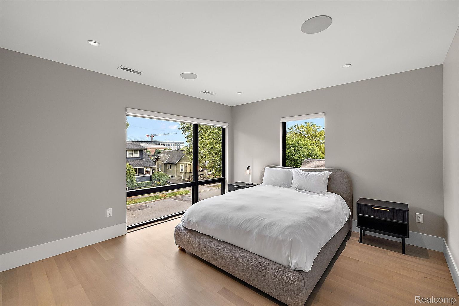This is a bright and modern primary bedroom featuring a large bed with white linens, flanked by dark nightstands. Large windows offer views of the neighborhood, and the light hardwood floors add warmth to the space. The room is painted in a neutral gray tone, creating a serene and inviting atmosphere.