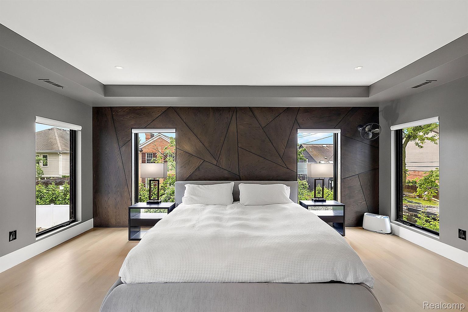 This is a primary bedroom featuring a modern design with a dark wood accent wall behind the bed. The room has light wood floors, gray walls, and two windows on either side of the bed, providing natural light. The bed is neatly made with white linens and gray bedding, and there are matching nightstands with lamps on either side.
