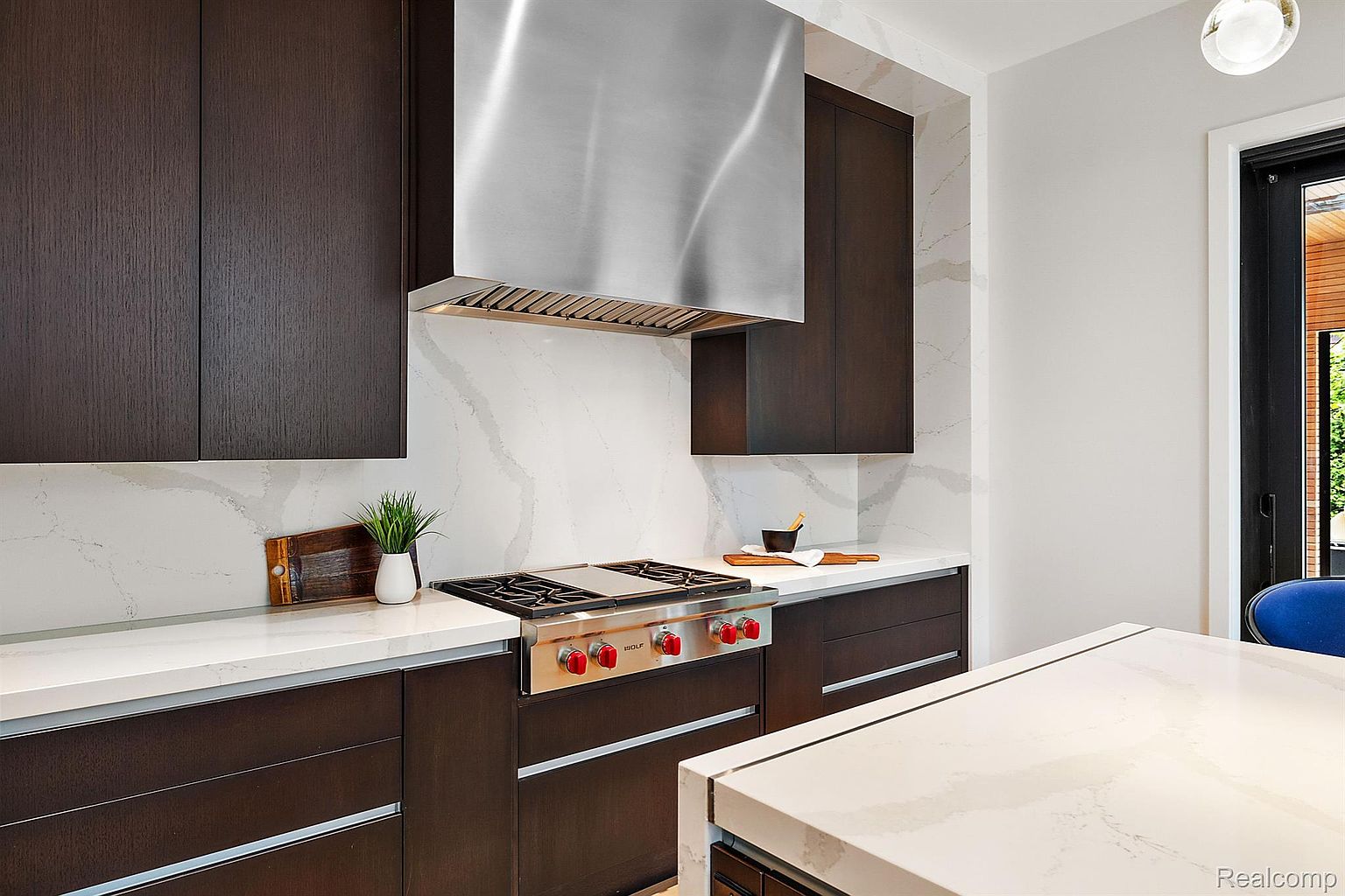 This is a modern kitchen featuring dark wood cabinetry, stainless steel appliances, and white quartz countertops. A stainless steel range hood is above the stove. The kitchen island is partially visible, and a window provides natural light.