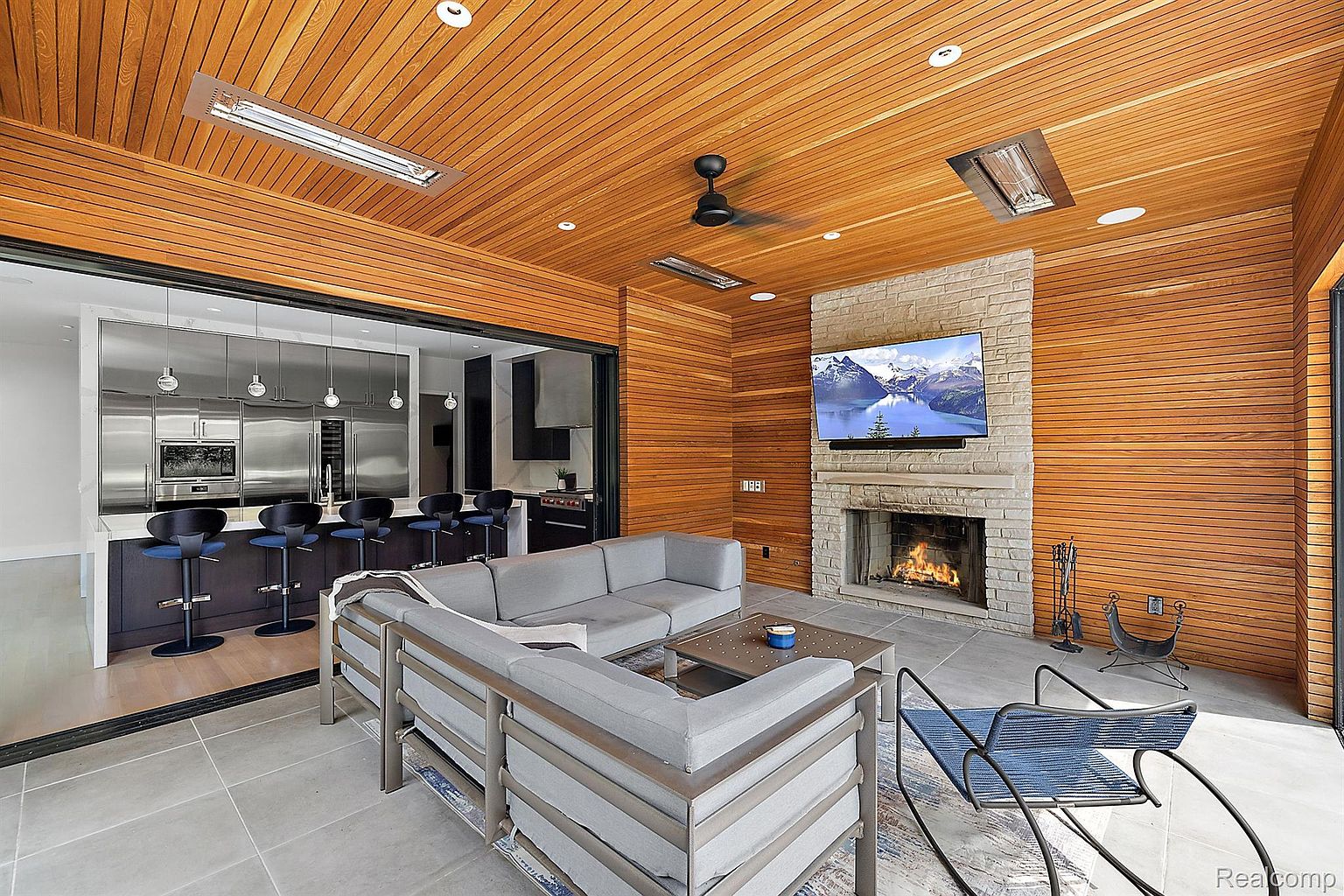 This is an indoor living space featuring a modern fireplace with a large television mounted above it. The room has a wood-paneled ceiling and walls, with a gray sectional sofa and a metal-framed chair providing seating. A sliding glass door opens to an adjacent kitchen area, creating an open and airy feel.