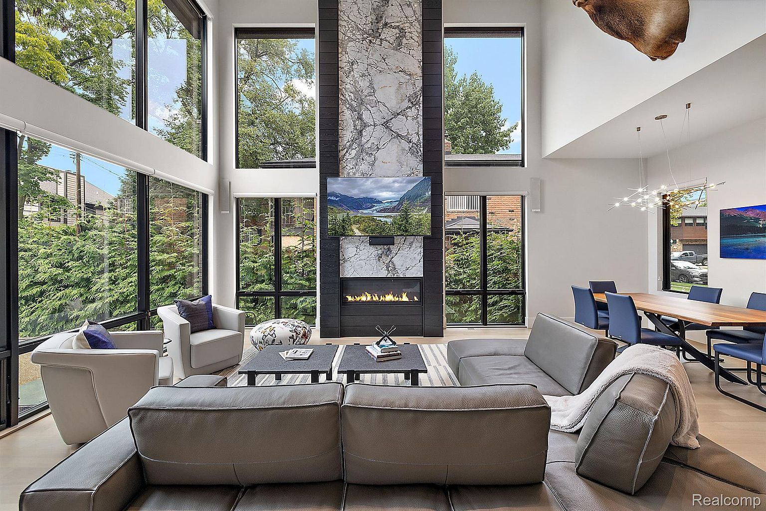 This is a spacious living room with high ceilings and large windows that offer ample natural light and views of the surrounding greenery. The room features a modern fireplace with a television mounted above it, a comfortable gray sectional sofa, and stylish armchairs. The open floor plan extends to a dining area, creating a seamless and inviting living space.