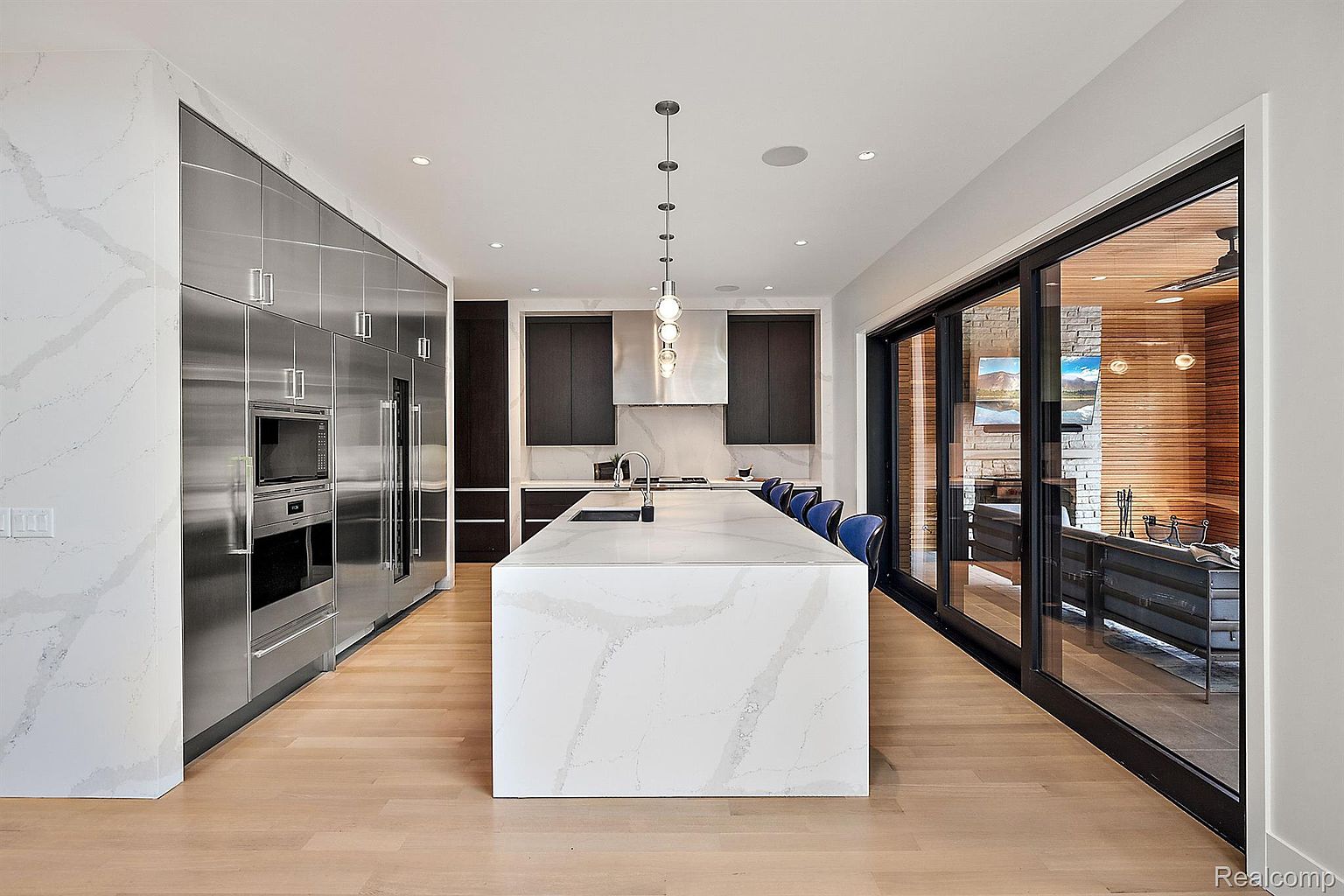 This is a bright and modern kitchen featuring a large island with a white marble countertop and a sink. Stainless steel appliances line one wall, while dark cabinets are featured on another. Sliding glass doors lead to an outdoor living space, creating an open and airy feel.