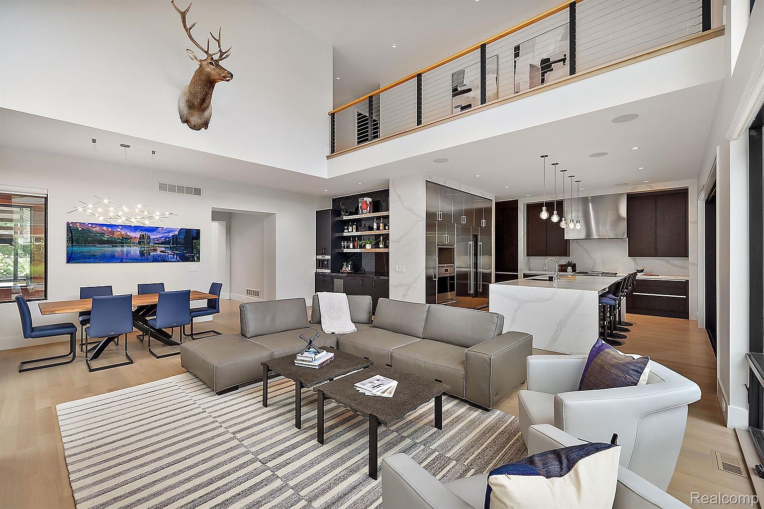 This is a spacious and modern living room featuring a large sectional sofa, two armchairs, and a striped area rug. The room has a high ceiling with a mounted deer head, a dining area with a large artwork, and an open-concept kitchen with a marble island. The overall impression is luxurious and contemporary, perfect for entertaining.