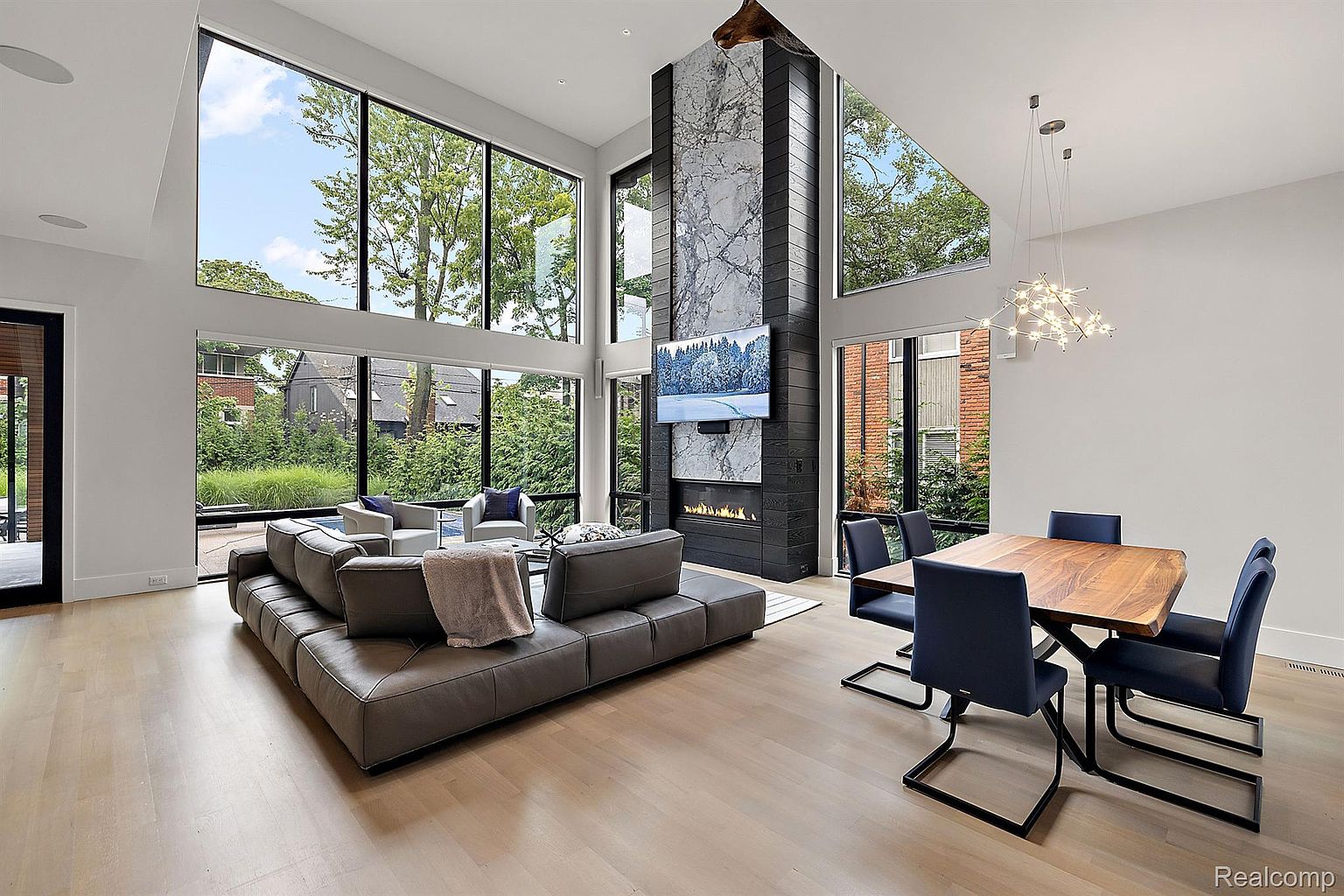 This is a spacious and modern living room with high ceilings and large windows offering ample natural light and views of the surrounding greenery. The room features a large sectional sofa, a fireplace with a marble surround and a mounted television, and a dining area with a wooden table and blue chairs. The overall impression is one of luxury and contemporary design.