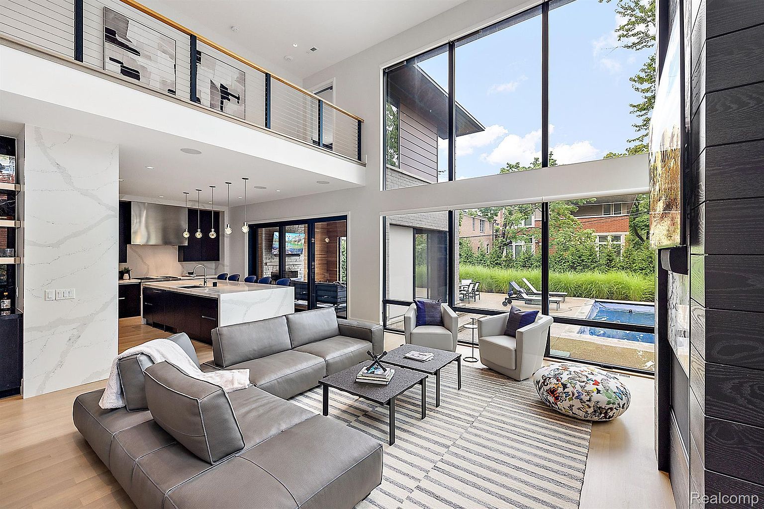 This is a spacious, modern living room with high ceilings and large windows offering a view of the backyard and pool. The room features a gray sectional sofa, two armchairs, and a striped rug. The open floor plan connects the living room to the kitchen, creating a seamless and inviting space.