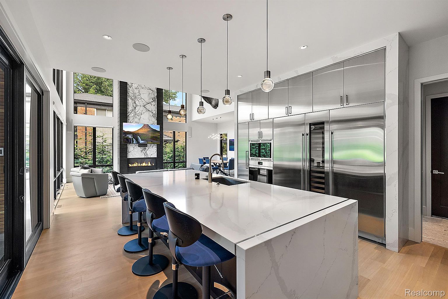 This is a bright, modern kitchen featuring a large island with a white countertop and blue bar stools. Stainless steel appliances line one wall, and a large window provides natural light and a view of the outdoors. The open floor plan connects the kitchen to the living area, creating a spacious and inviting atmosphere.