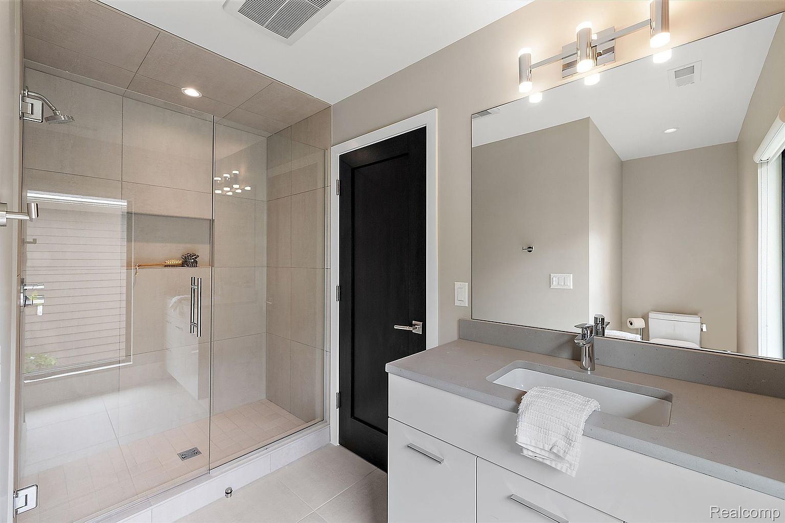 This is a modern primary bathroom featuring a glass-enclosed shower with tile walls and a built-in niche. The vanity has a gray countertop, white cabinets, and a rectangular sink with a chrome faucet. A large mirror with modern lighting fixtures hangs above the vanity, and a black door adds a touch of contrast to the neutral color palette.