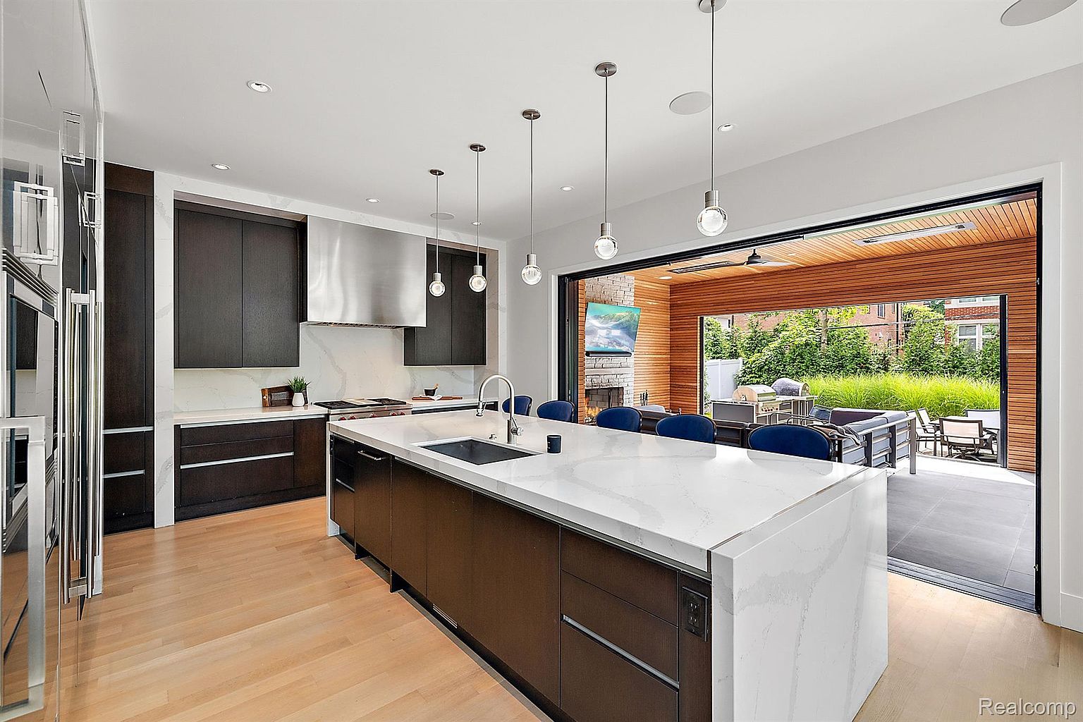 This is a modern kitchen featuring dark wood cabinetry, a large white countertop island with a sink, and stainless steel appliances. Pendant lights hang above the island, and a large sliding glass door opens to an outdoor patio area with lounge furniture and a fireplace. The kitchen has a clean, contemporary design with an open layout.