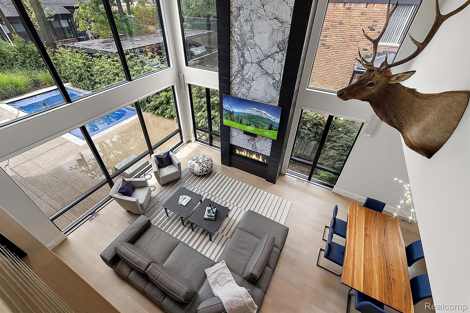 This is an interior shot of a modern living room with floor-to-ceiling windows offering views of the exterior. The room features a gray sectional sofa, two armchairs, a striped area rug, and a fireplace with a mounted television. A dining area with a wooden table and blue chairs is also visible, along with a mounted deer head, creating a luxurious and spacious living area.