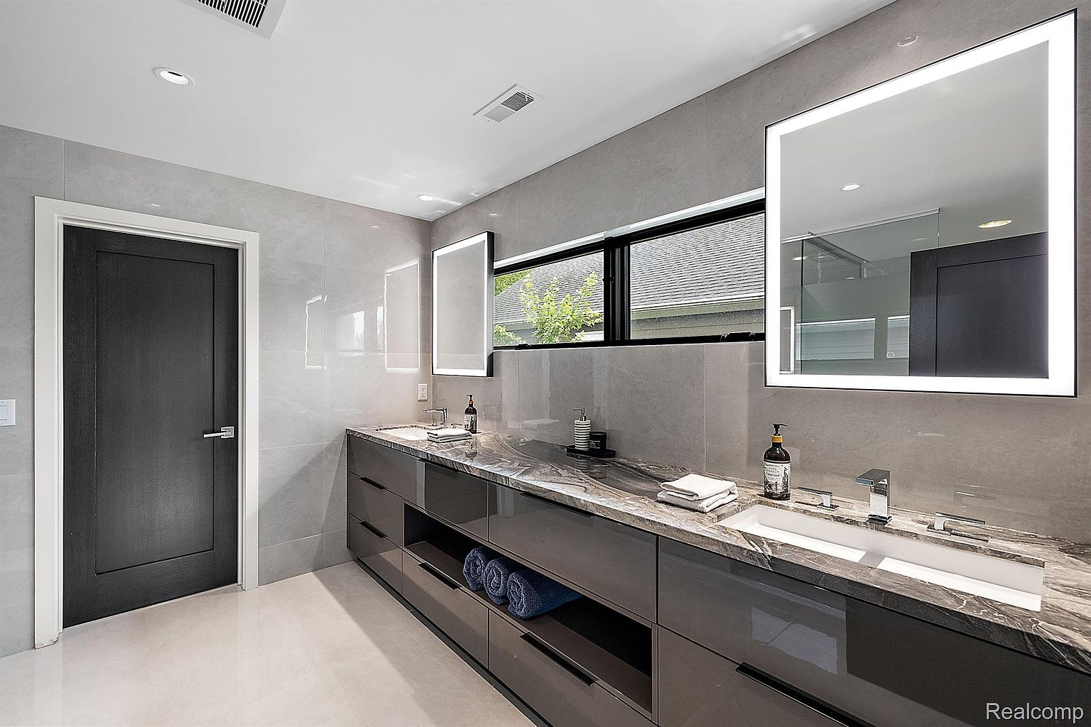 This is a modern primary bathroom featuring a double vanity with a marble countertop and sleek, dark gray cabinetry. Two illuminated mirrors hang above the sinks, and a window provides natural light. The walls are covered in large gray tiles, and the floor is a light neutral color, creating a clean and luxurious aesthetic.