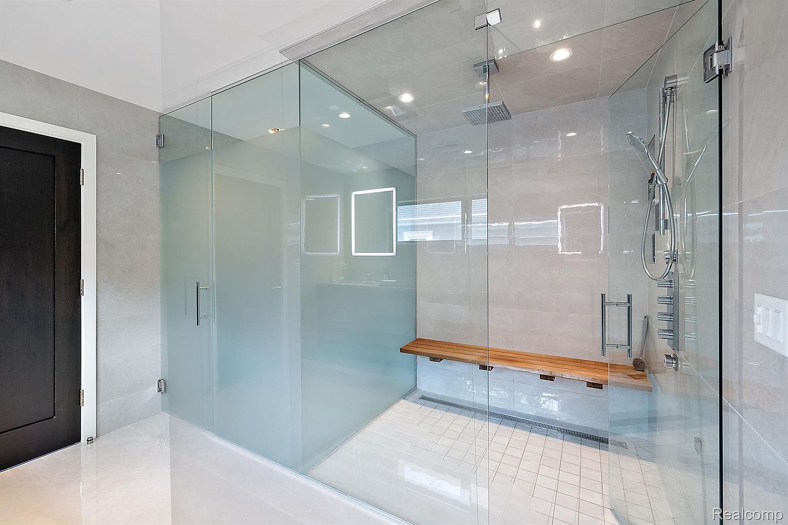 This is a modern primary bathroom featuring a glass-enclosed shower with a built-in wooden bench and a rainfall showerhead. The walls are tiled in a light gray, and the floor is white tile. The shower has a sleek, minimalist design with a frameless glass door and a hand-held shower attachment.