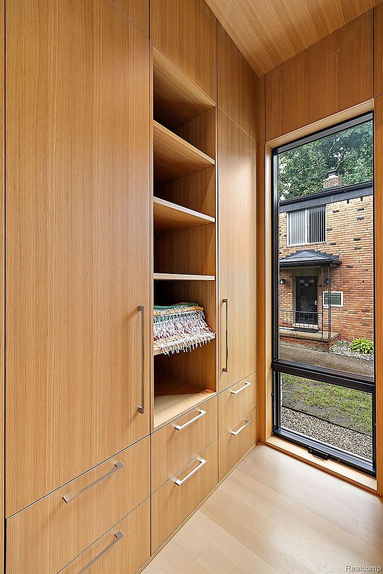 This image showcases a modern walk-in closet with light wood cabinetry, featuring both closed storage with sleek, silver handles and open shelving. A window provides natural light, offering a view of a brick building outside. The closet's design is clean and minimalist, emphasizing functionality and style.