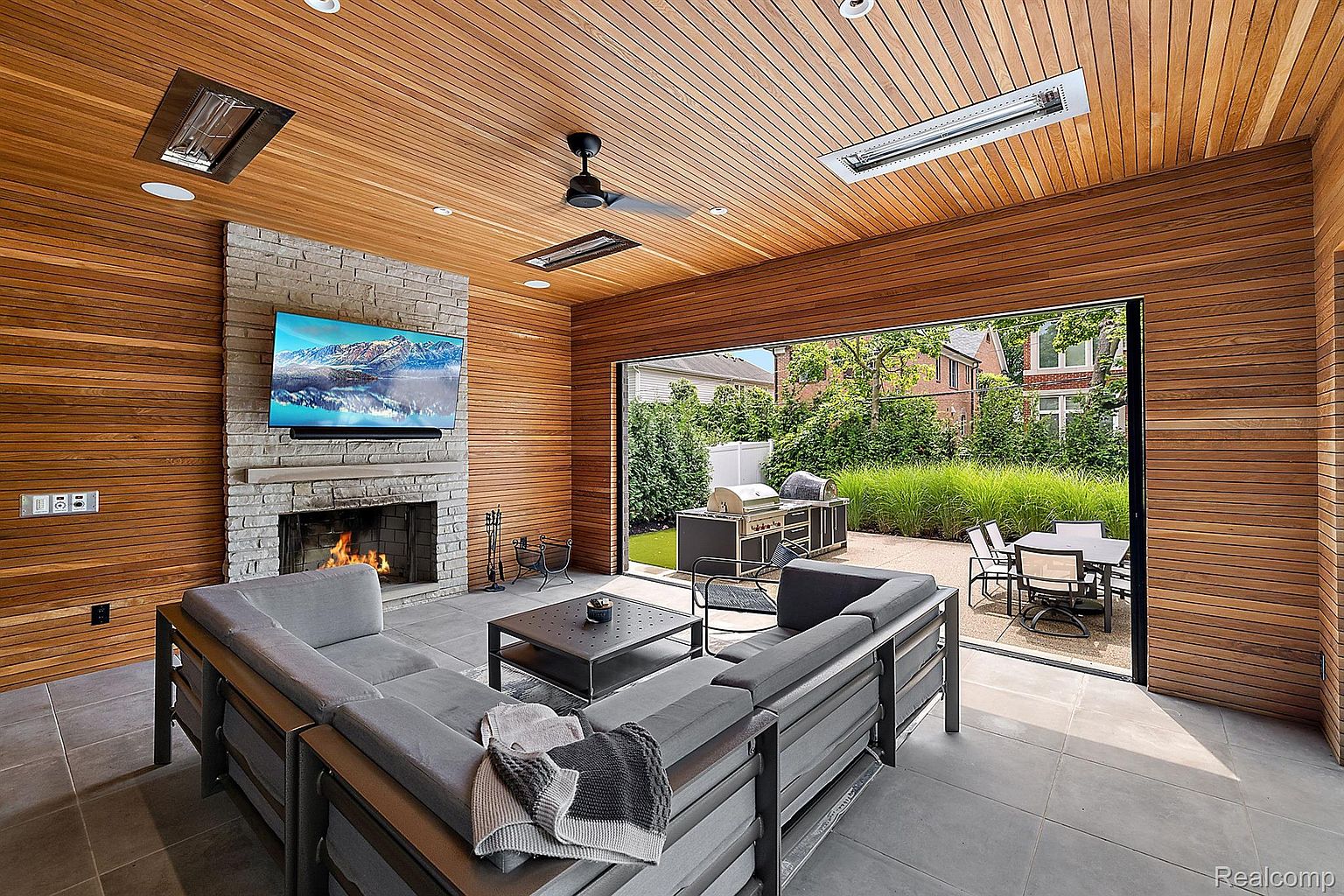 This image showcases a modern sunroom with wood-paneled walls and ceiling, featuring a stone fireplace with a mounted TV. A large sliding glass door opens to an outdoor patio with a grill and dining area. The room is furnished with a gray sectional sofa and a dark coffee table, creating a seamless indoor-outdoor living space.