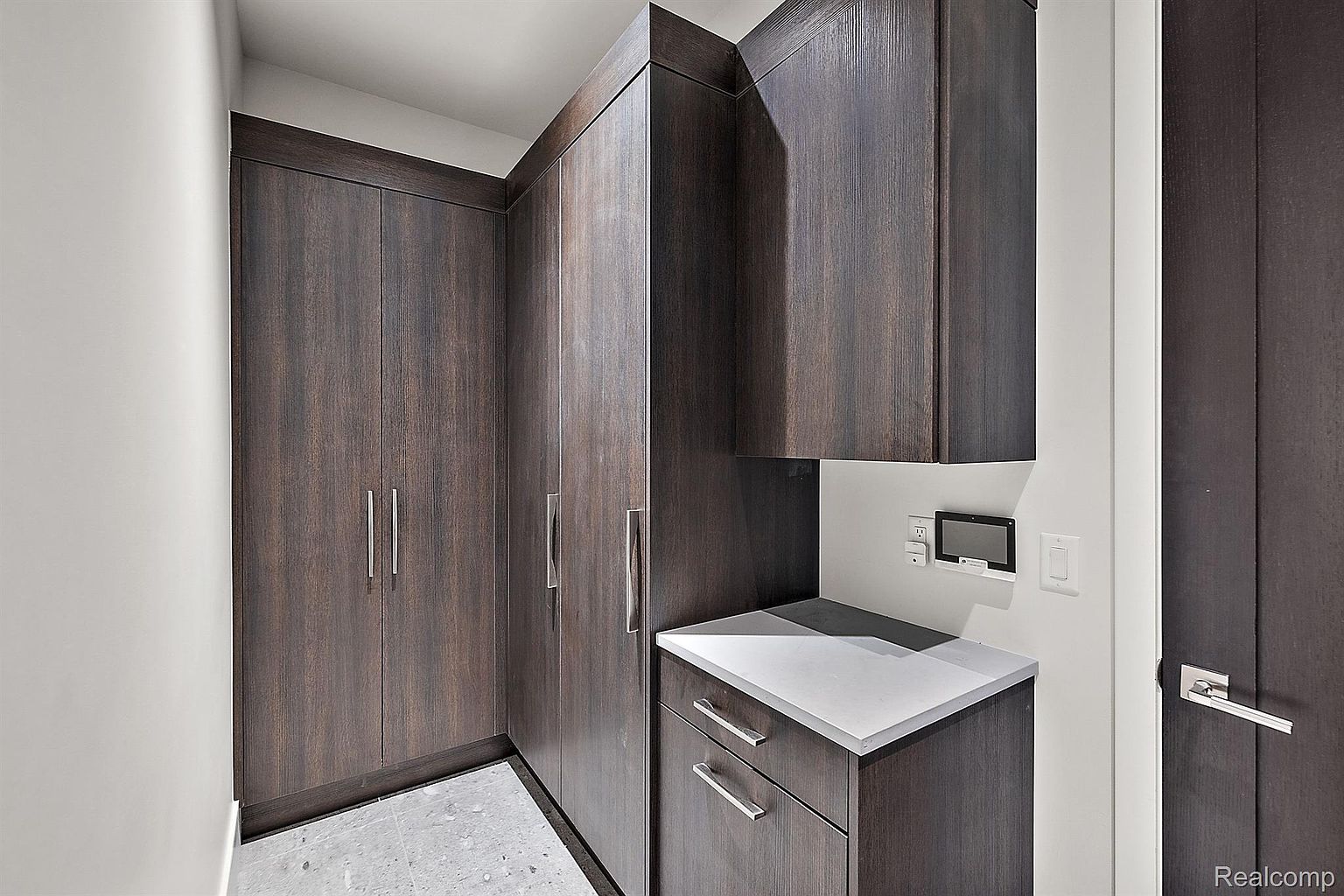 This interior shot showcases a modern laundry room with dark wood cabinetry and sleek silver hardware. A light countertop provides workspace, and a control panel is mounted on the wall. The room appears well-organized and functional, ideal for a contemporary home.