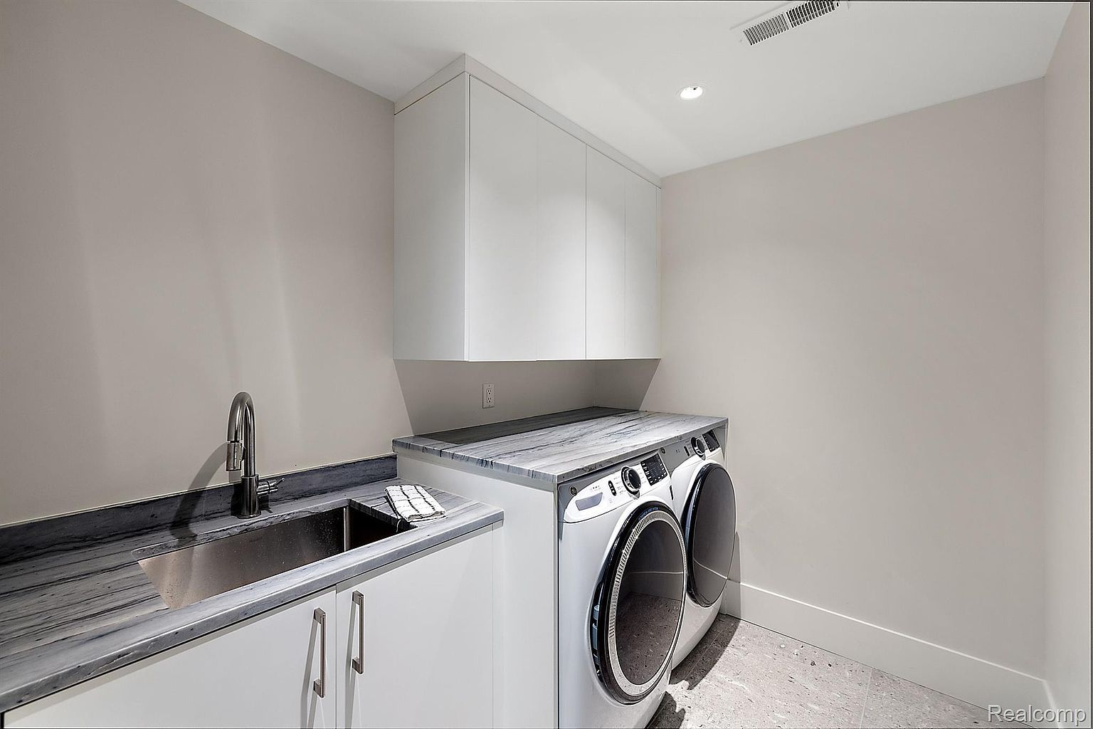 This is a modern laundry room featuring a front-loading washer and dryer set with a countertop above. The room includes a stainless steel sink with a modern faucet, white cabinets, and a gray countertop that matches the sink area. The overall design is clean and functional, with a focus on maximizing space and providing a stylish laundry area.