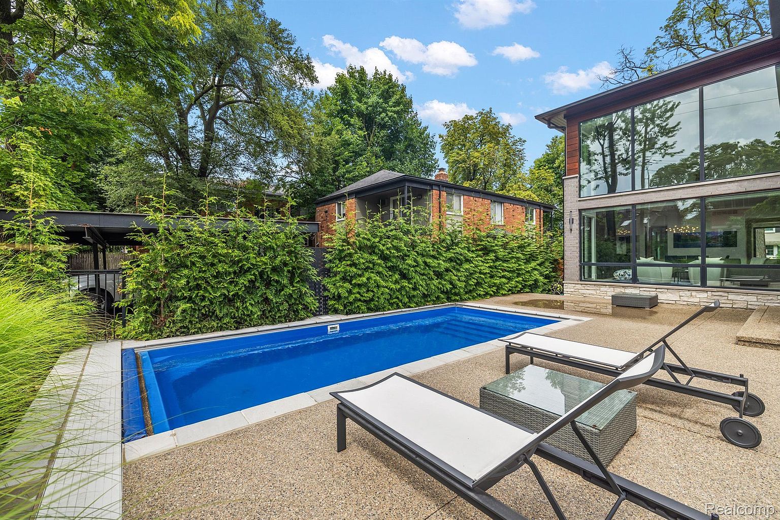 This image showcases a luxurious backyard pool area, featuring a rectangular pool with clear blue water, surrounded by a concrete patio. Two modern lounge chairs and a small table are positioned near the pool, inviting relaxation. The backdrop includes lush greenery, a brick house, and a modern house with large windows, creating a private and upscale outdoor living space.