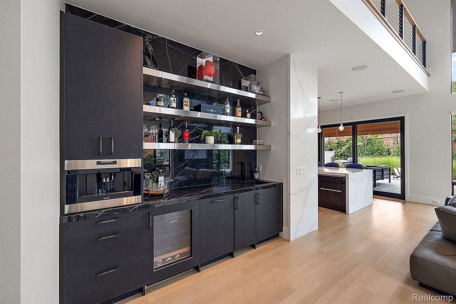 This interior shot showcases a modern kitchen area with dark cabinetry, marble countertops and backsplash, and stainless steel appliances, including a built-in coffee machine and wine cooler. Open shelving displays glassware and decorative items, adding a touch of elegance. The space flows into a dining area with a view of the outdoors, creating an inviting and sophisticated atmosphere.