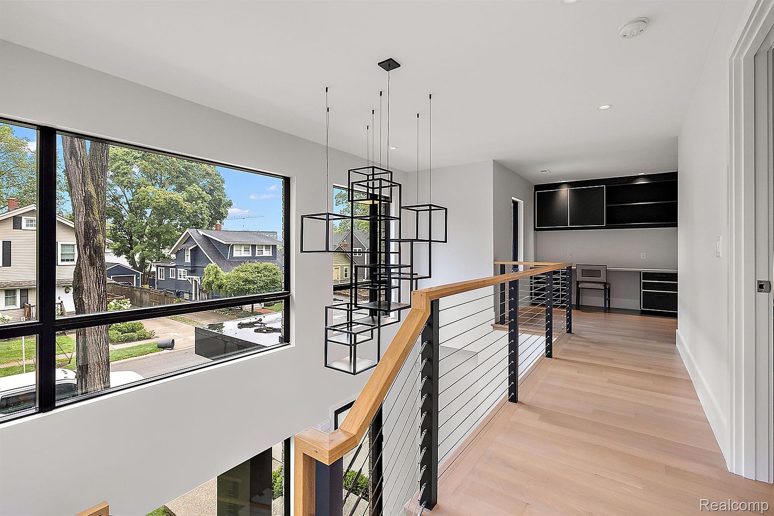 This is an interior shot of a modern hallway and staircase area. The space features a large window with a view of the neighborhood, a contemporary chandelier made of geometric shapes, and a railing with wooden accents and metal cables. An office area is visible in the background, suggesting a functional and stylish design.