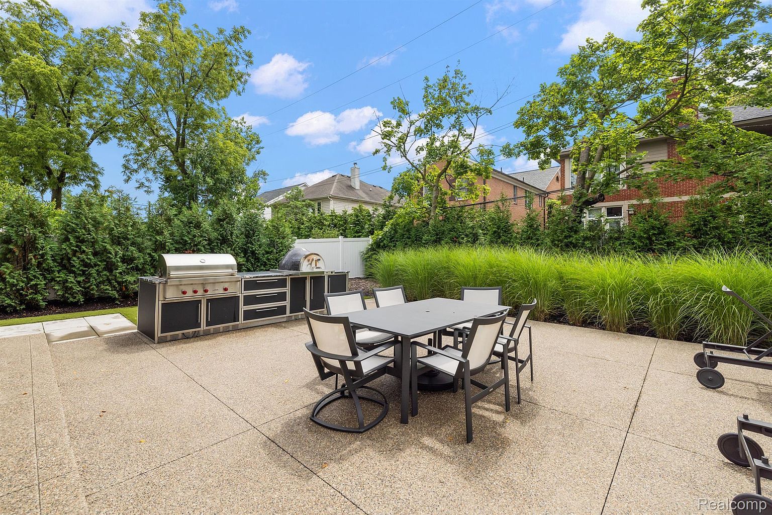 This image showcases a well-appointed outdoor patio area, perfect for entertaining. A modern outdoor dining set with seating for six is centrally located, complemented by a built-in outdoor kitchen featuring a stainless steel grill and ample counter space. Lush greenery surrounds the patio, providing privacy and a serene atmosphere.