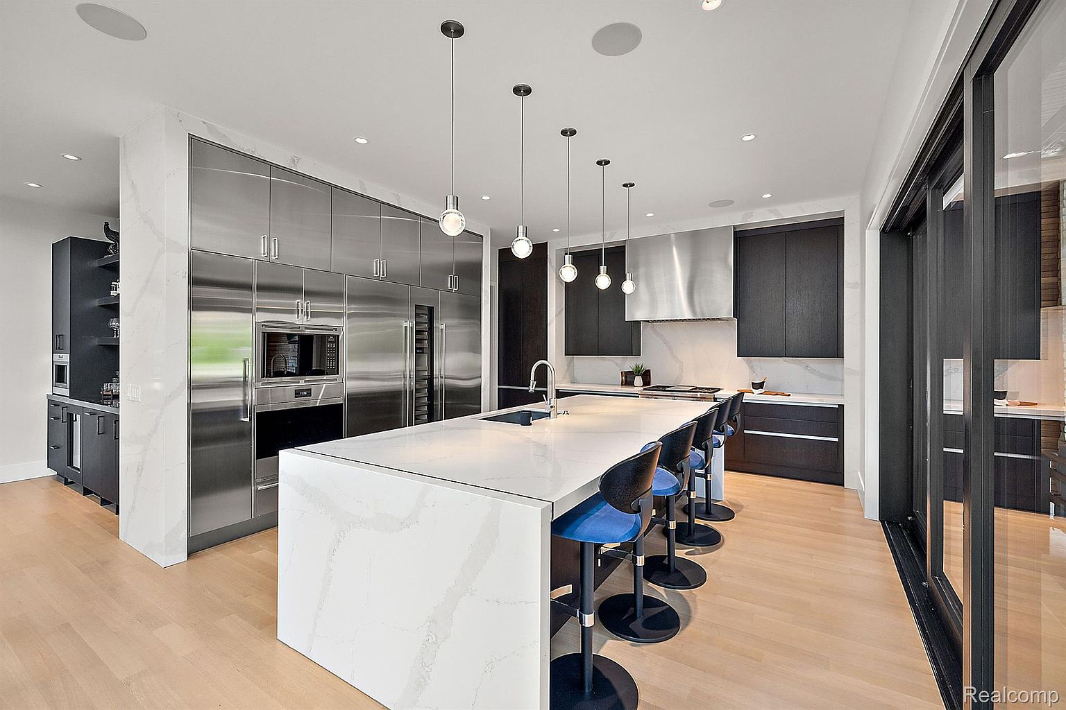 This is a modern kitchen with stainless steel appliances, including a large refrigerator and built-in oven. A large white island with a waterfall edge provides ample counter space and seating with blue bar stools. Pendant lights hang above the island, and dark cabinetry complements the stainless steel and white finishes, creating a sleek and contemporary aesthetic.