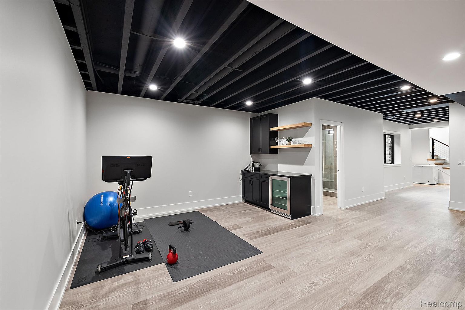 This is a well-lit home gym featuring a stationary bike, exercise ball, and free weights on a black mat. The room includes a built-in bar area with cabinets, open shelving, and a beverage cooler. The ceiling is an exposed black beam design, and the flooring is light-colored wood, creating a modern and functional workout space.