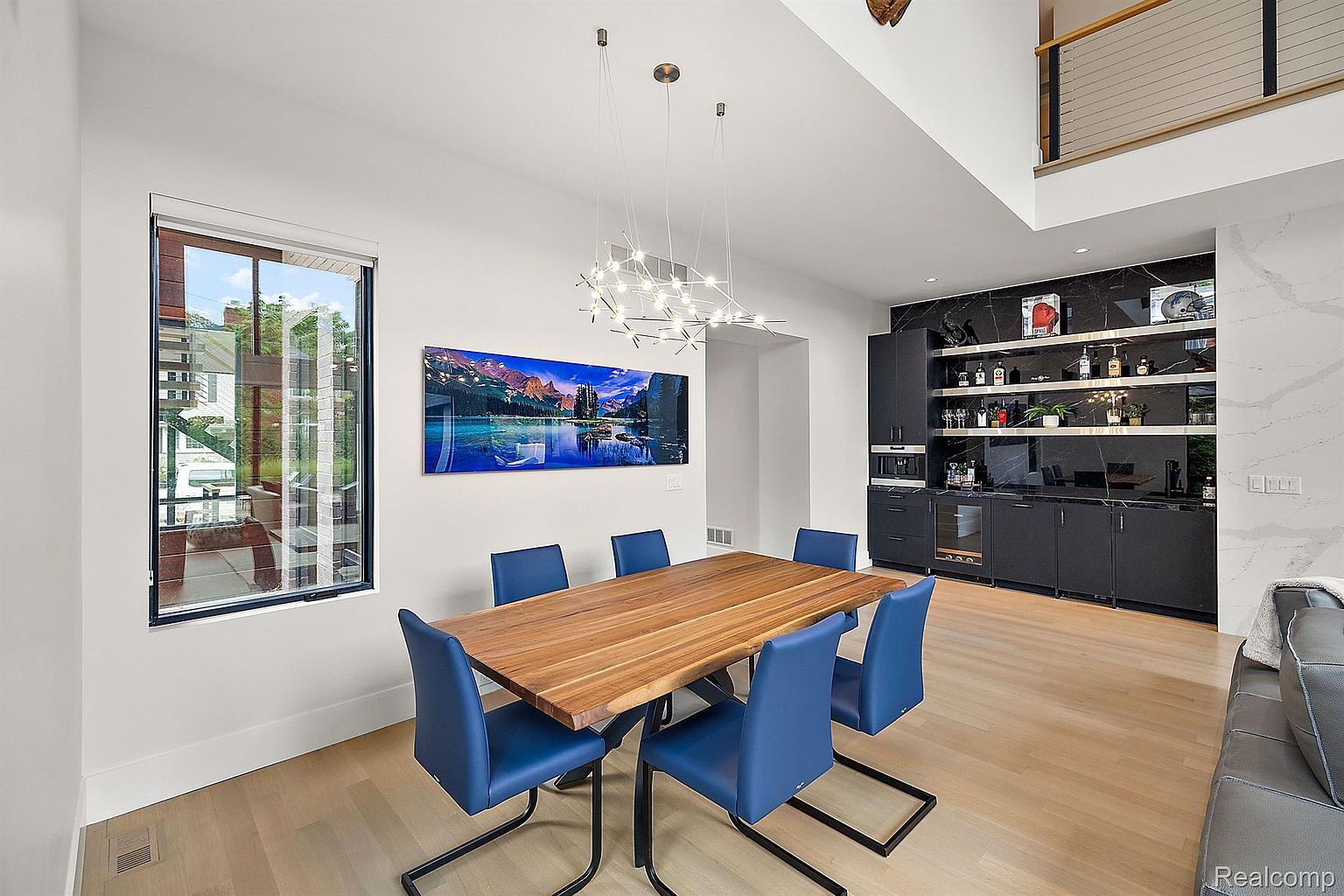 This is a dining room featuring a wooden table with blue chairs, set against a backdrop of a modern bar area with dark cabinetry and marble accents. A large window provides natural light, and a contemporary chandelier hangs above the table. The room exudes a sophisticated and stylish ambiance, perfect for both casual meals and formal gatherings.