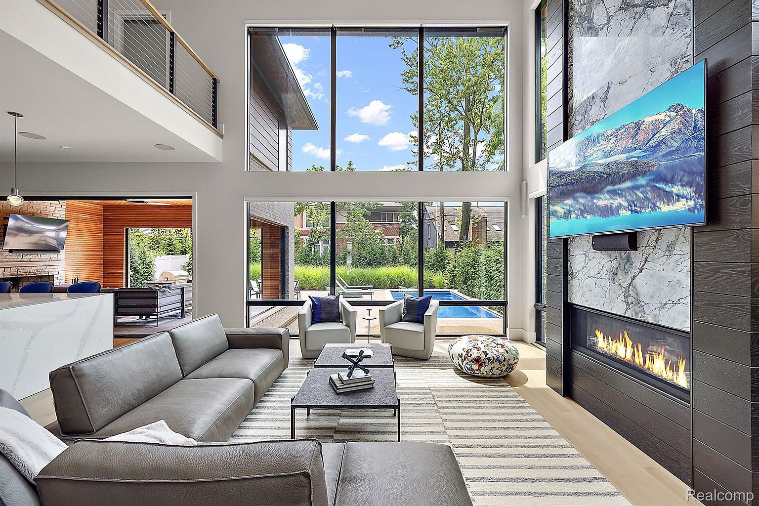 This is a spacious and modern living room with high ceilings and large windows offering views of the outdoor pool and greenery. The room features a gray sectional sofa, two armchairs, a striped rug, and a fireplace with a marble surround and a mounted television. The overall design is contemporary and luxurious, creating a comfortable and stylish living space.
