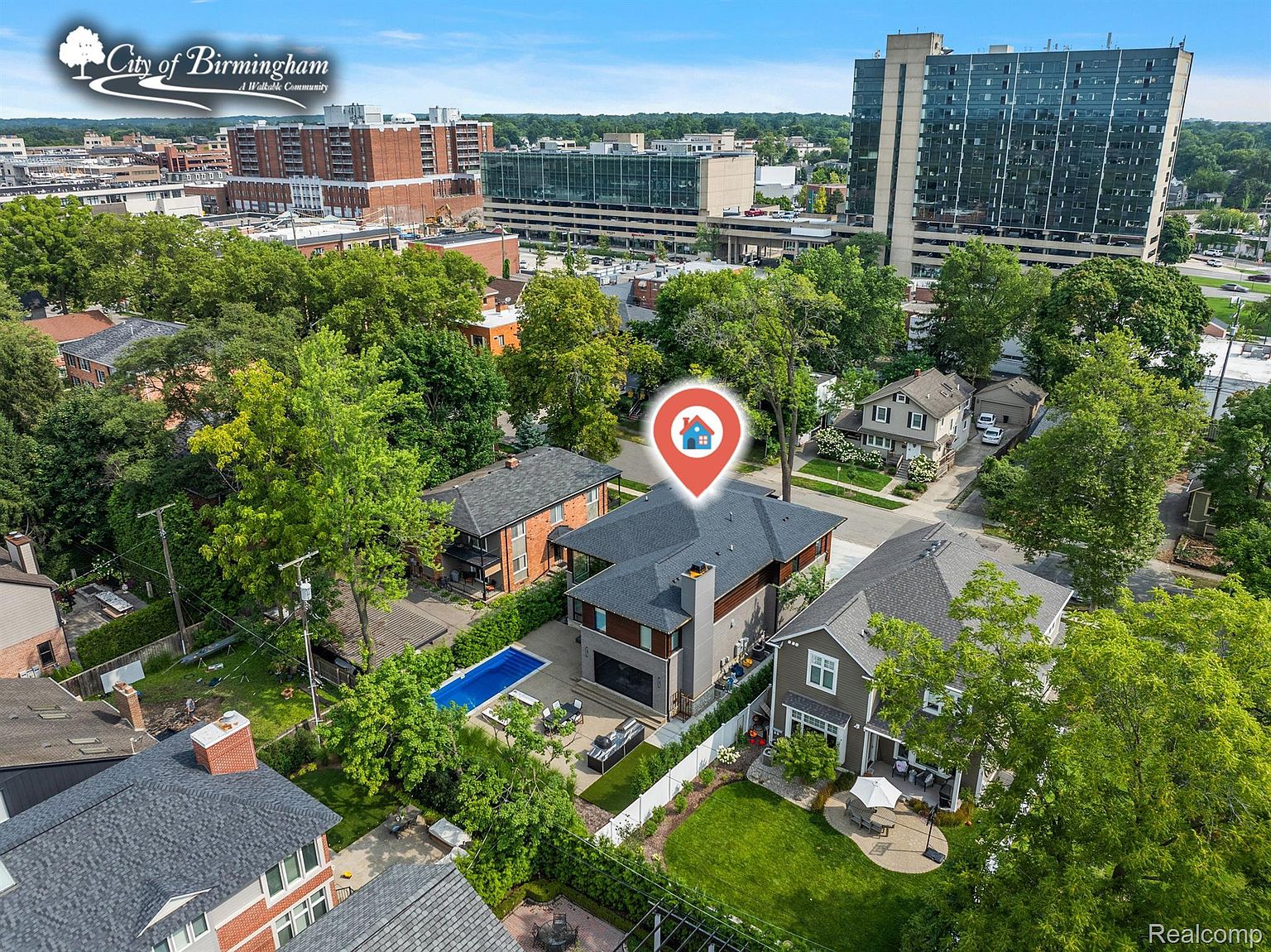 This aerial view showcases a residential property nestled among lush greenery in an urban setting. The house features a modern design with a dark roof and a backyard pool, complemented by a well-manicured lawn and patio area. The surrounding neighborhood includes a mix of residential buildings and high-rise structures, creating a vibrant and desirable location.
