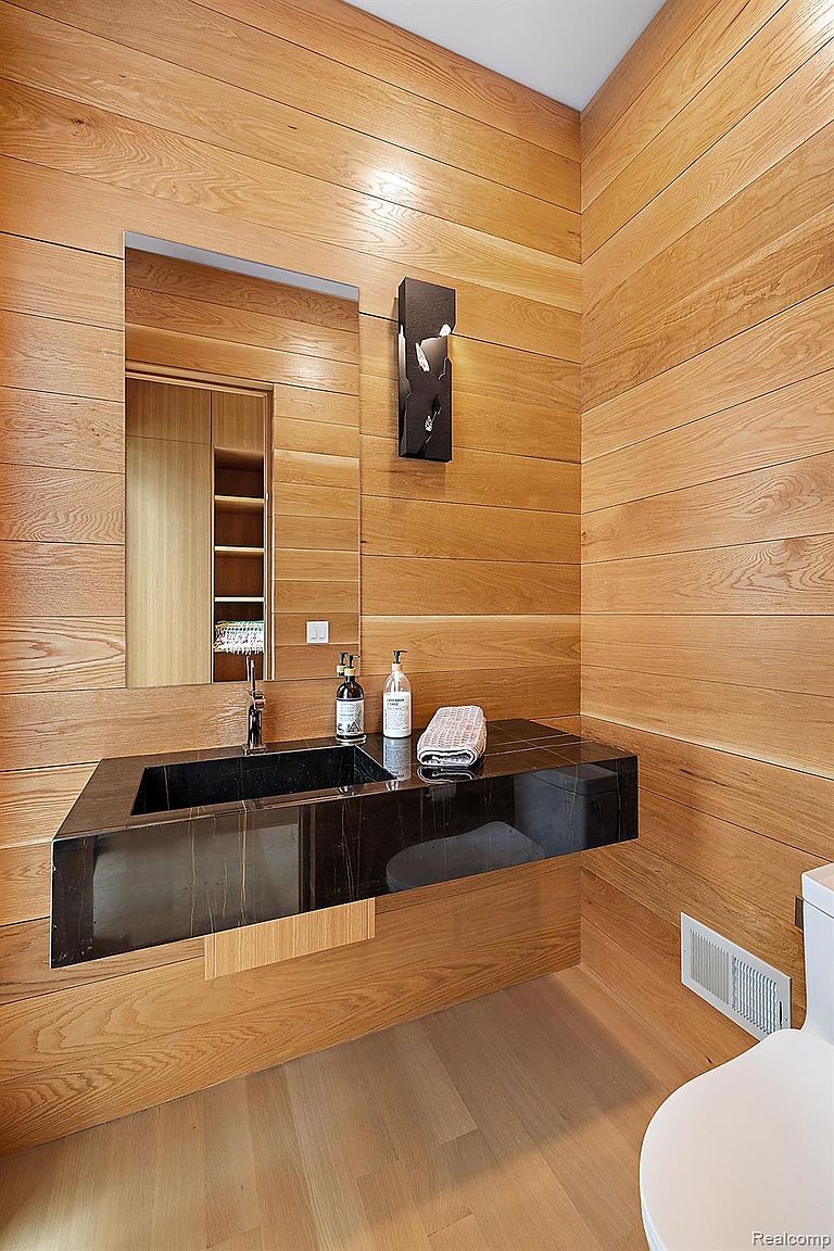 This is a modern bathroom featuring wood paneled walls and a black marble countertop with an integrated sink. A large mirror reflects a built-in shelving unit, adding depth to the space. The overall design is sleek and minimalist, creating a spa-like atmosphere.