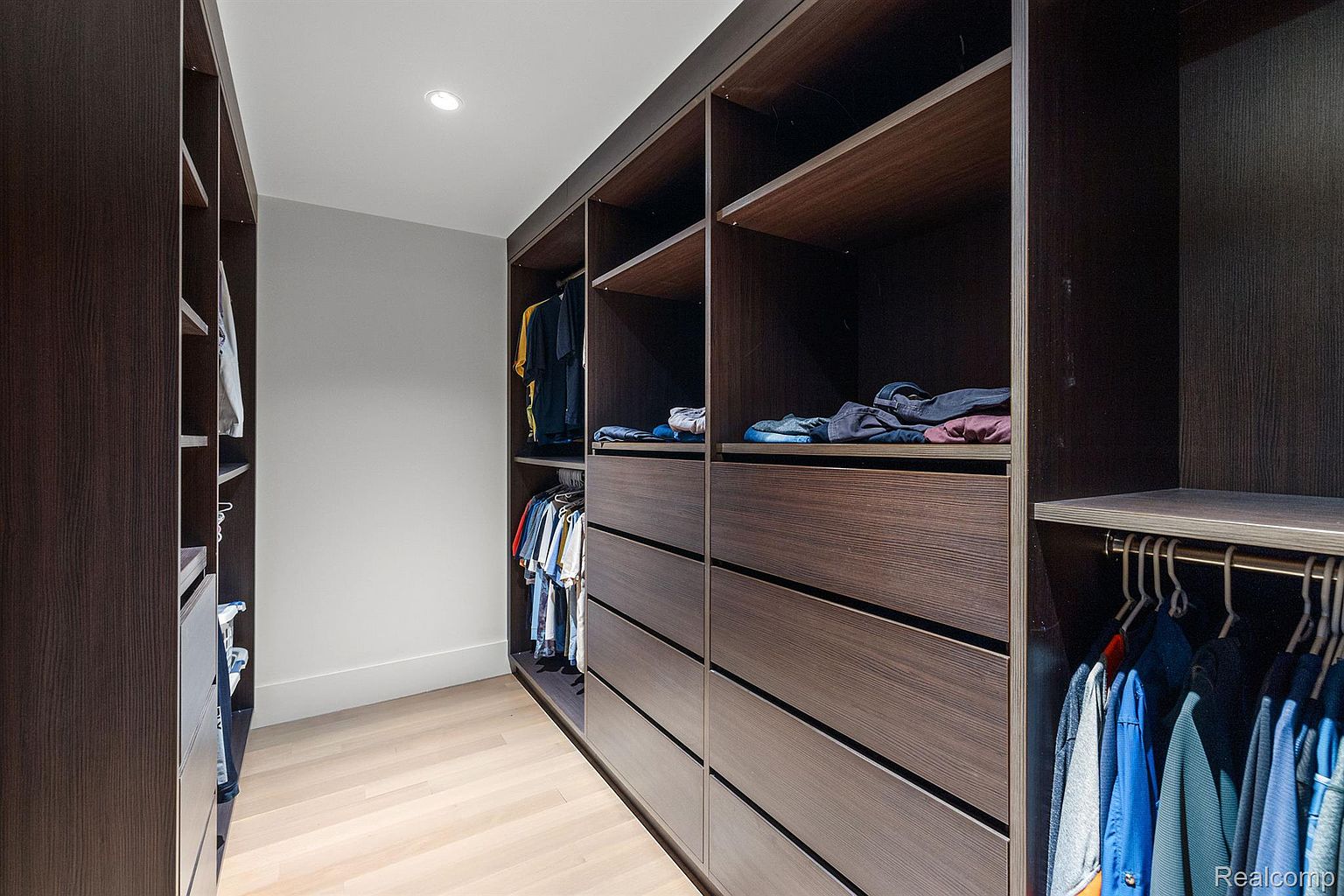 This is a well-organized walk-in closet featuring dark wood shelving and drawers. Clothes are neatly hung and folded, maximizing the storage space. The light wood flooring and recessed lighting create a clean and modern aesthetic, perfect for showcasing the closet's functionality and style.