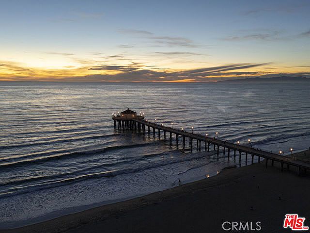 This aerial perspective captures a serene sunset over the ocean, highlighting a long wooden pier extending from the sandy beach into the water. The pier features a central structure at its end and is illuminated by a series of warm lights that create a welcoming path across the dark sea. The composition emphasizes the vast horizon and the tranquil atmosphere of the coastal setting.