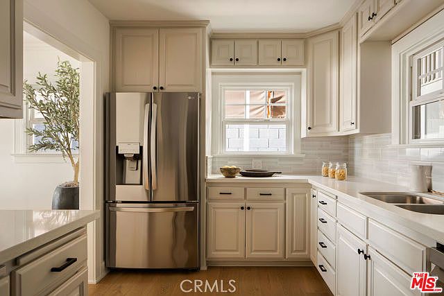 This bright and clean kitchen features light-toned cabinetry, a stainless steel refrigerator, and a double-basin sink set into a light-colored countertop. A window above the sink provides natural light, while a subway-style tile backsplash adds a classic touch to the space. The perspective is a straight-on view of the kitchen work area, highlighting the efficient layout and neutral color palette.