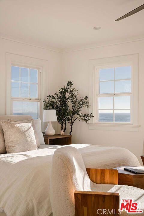 This serene bedroom features a neutral color palette and a cozy, textured aesthetic, centered around a plush bed with layered linens. Two large windows frame a stunning, unobstructed view of the ocean, filling the room with natural light. A decorative potted plant and a minimalist bedside lamp add a touch of organic warmth to the space, creating a tranquil and inviting atmosphere.
