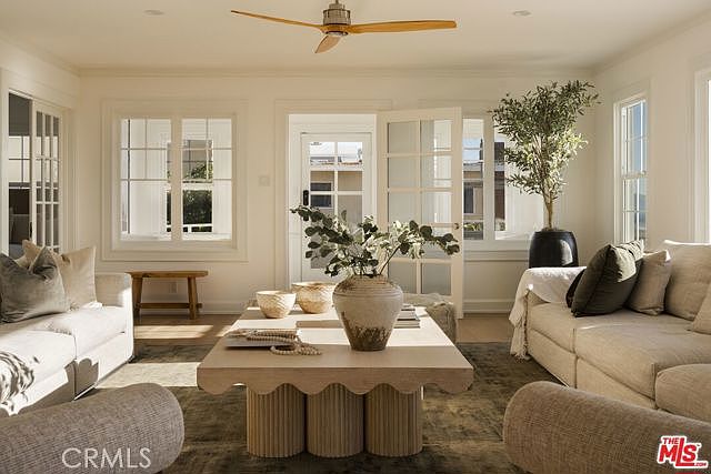 This bright and airy living room features a neutral color palette with plush, light-toned sofas arranged around a unique, scalloped-base coffee table. Large windows and glass-paned doors allow for abundant natural light, creating a seamless connection to the outdoors. A ceiling fan with light wood blades centers the room, while a potted olive tree adds a touch of organic greenery to the sophisticated, contemporary space.