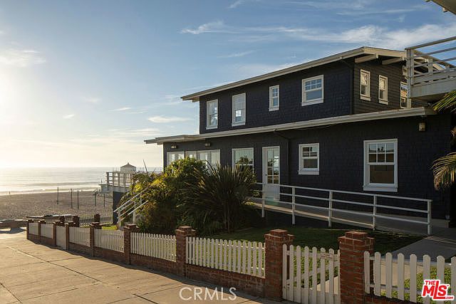 This side view captures a dark-sided, multi-story coastal home situated directly adjacent to a sandy beach. The property features a white picket fence with brick pillars, a ramp leading to the entrance, and numerous windows that offer views of the ocean. The bright, sunny lighting highlights the home's classic architectural style and its prime beachfront location.