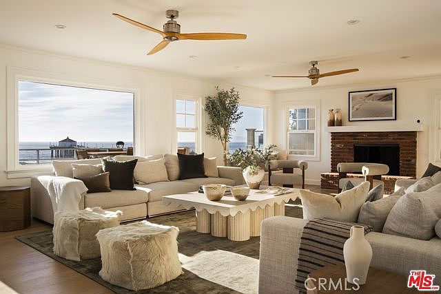 This bright and airy living room features a neutral color palette with plush seating, including a large sectional sofa and two textured ottomans. A unique, scalloped-base coffee table sits at the center of the space, while a brick-clad chimney breast anchors the room near a cozy fireplace. Large windows provide expansive views of the ocean and a pier, creating a serene and sophisticated coastal atmosphere.