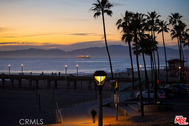 This scenic, wide-angle shot captures a serene beachside walkway at sunset, featuring a prominent street lamp in the foreground and a silhouette of a person walking along the path. The composition includes a wooden pier extending into the ocean, with cargo ships visible on the horizon against a vibrant, multi-colored sky. Tall palm trees frame the right side of the scene, creating a tranquil and picturesque coastal atmosphere.