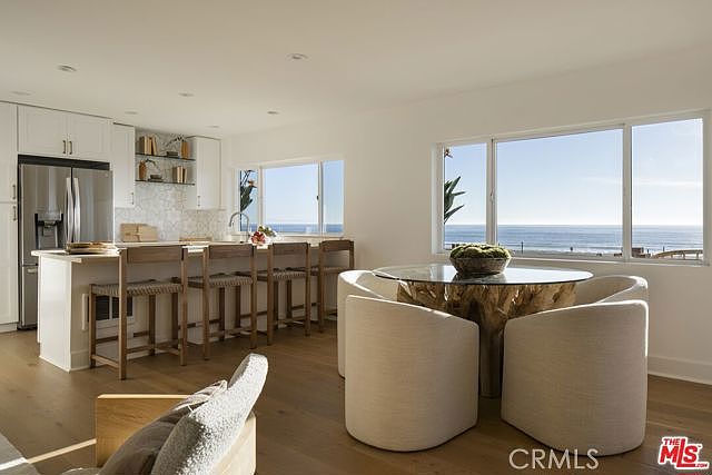 This bright, open-concept kitchen and dining area features light wood flooring and a clean, modern aesthetic. The kitchen includes a central island with bar seating, white cabinetry, and a patterned tile backsplash, while the adjacent dining space is anchored by a unique organic-shaped wooden table and curved chairs. Large windows provide an expansive, unobstructed view of the ocean, creating a serene and airy atmosphere.