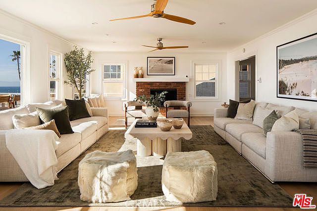 This bright and airy living room features a symmetrical layout with two large, light-colored sofas facing each other across a textured, scalloped-base coffee table. A brick fireplace serves as the central focal point, flanked by two armchairs and topped with a framed landscape print. The space is anchored by a large, muted-tone area rug and two plush, furry ottomans, creating a sophisticated yet comfortable coastal atmosphere with views of the ocean visible through the side windows.