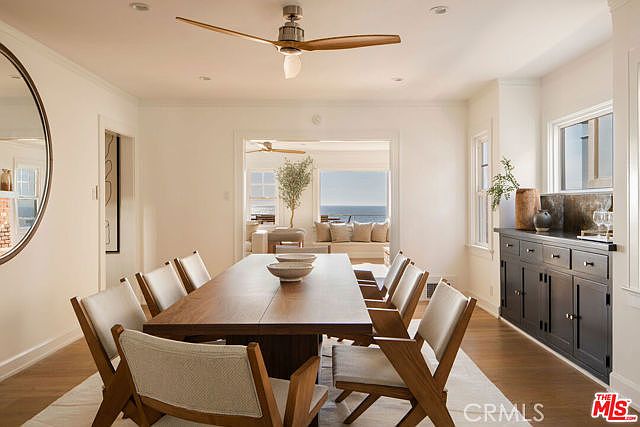This elegant dining room features a long wooden table surrounded by modern chairs with light-toned upholstery, set beneath a sleek ceiling fan. To the right, a dark-toned sideboard provides storage and display space, while a large circular mirror on the left wall adds depth to the room. The space opens up to a bright, sunlit living area with a view of the ocean, creating a seamless and airy coastal atmosphere.