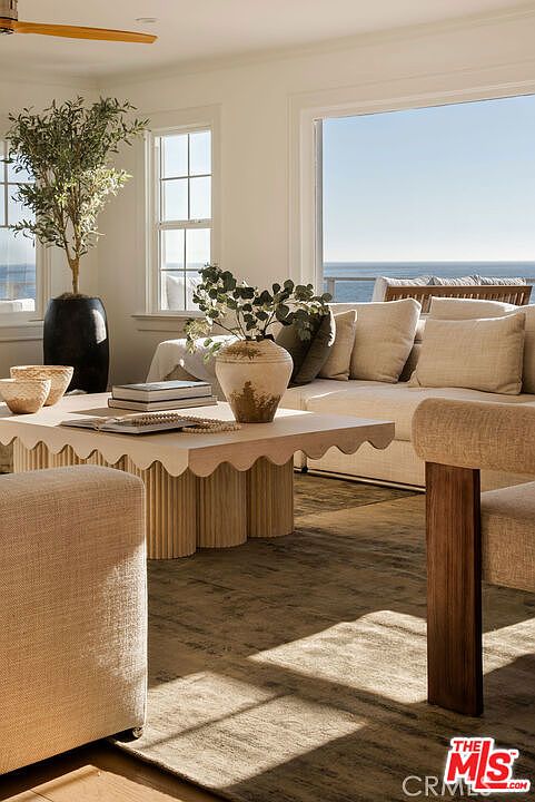 This bright and airy living room features a coastal-inspired aesthetic with a neutral color palette and soft textures. A unique, scalloped-edge coffee table with a fluted base serves as the centerpiece, complemented by plush seating and a large, textured area rug. Large windows provide an expansive, unobstructed view of the ocean, bathing the space in natural light.