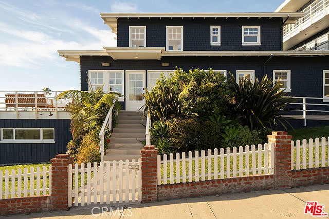 This charming two-story home features dark blue shingle siding and a classic architectural style with a prominent front entryway. A white picket fence with brick pillars lines the front of the property, leading to a set of stairs that ascend to the main entrance. Lush greenery and mature landscaping surround the front steps, while a raised deck area is visible on the left side of the structure.